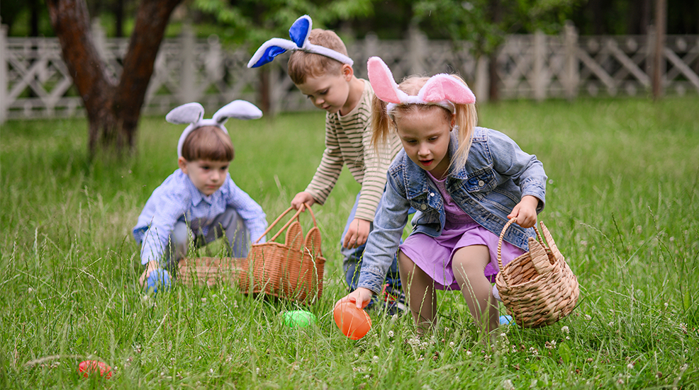 A group of young children with bunny ear headbands search for colorful Easter eggs in tall green grass, holding wicker baskets. This joyful outdoor scene captures the spirit of Easter traditions, springtime fun, childhood play, and festive seasonal activities.